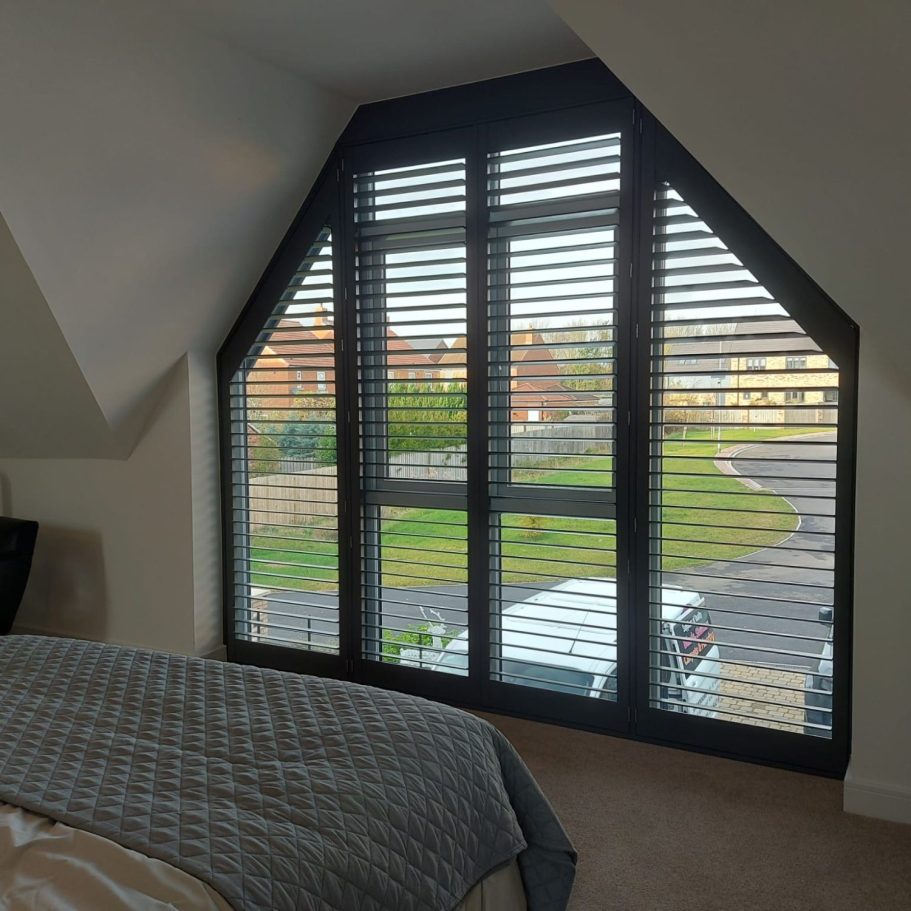 A cozy bedroom corner with large, angled windows and blinds overlooking a garden.