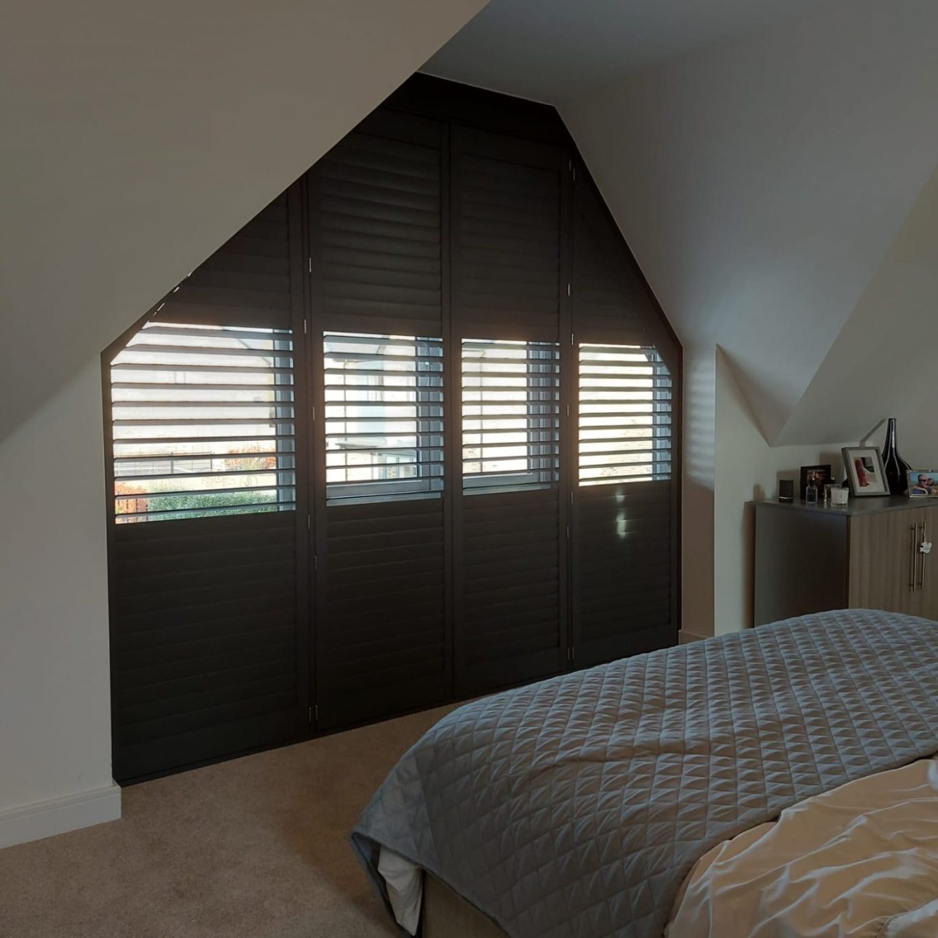 Shutters A bedroom with sloped walls, a bed, and dark wooden shutters on a window.