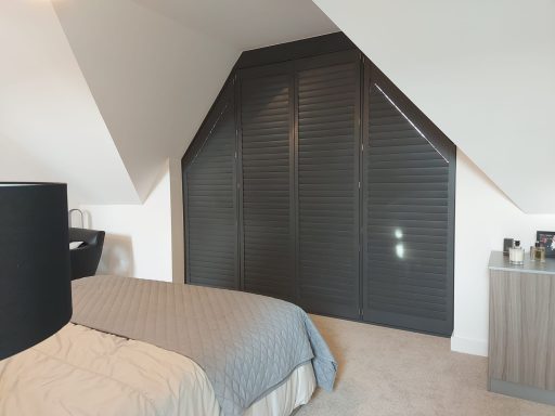 A modern bedroom with slanted wall, bed, and dark blinds covering a large window.