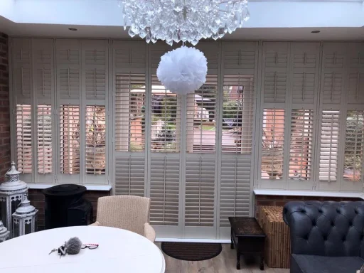 A room with wooden shutters, a chandelier, and a round table with plush seating.