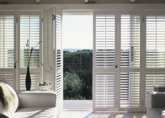 Bright room with white blinds, a sleek vase, and a view of greenery outside.