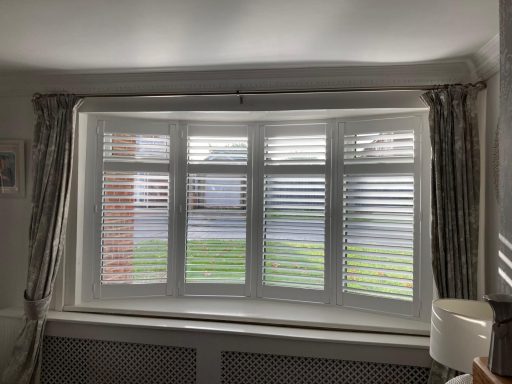 Bay window with white blinds and grey curtains, revealing green grass outside.