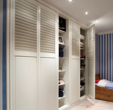 White wardrobe with shutter doors and shelves, beside a bed in a striped room.