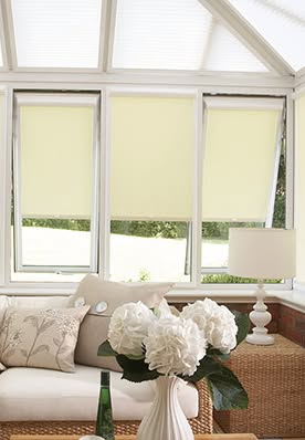 A bright conservatory with yellow blinds, white flowers, and a lamp on a woven table.