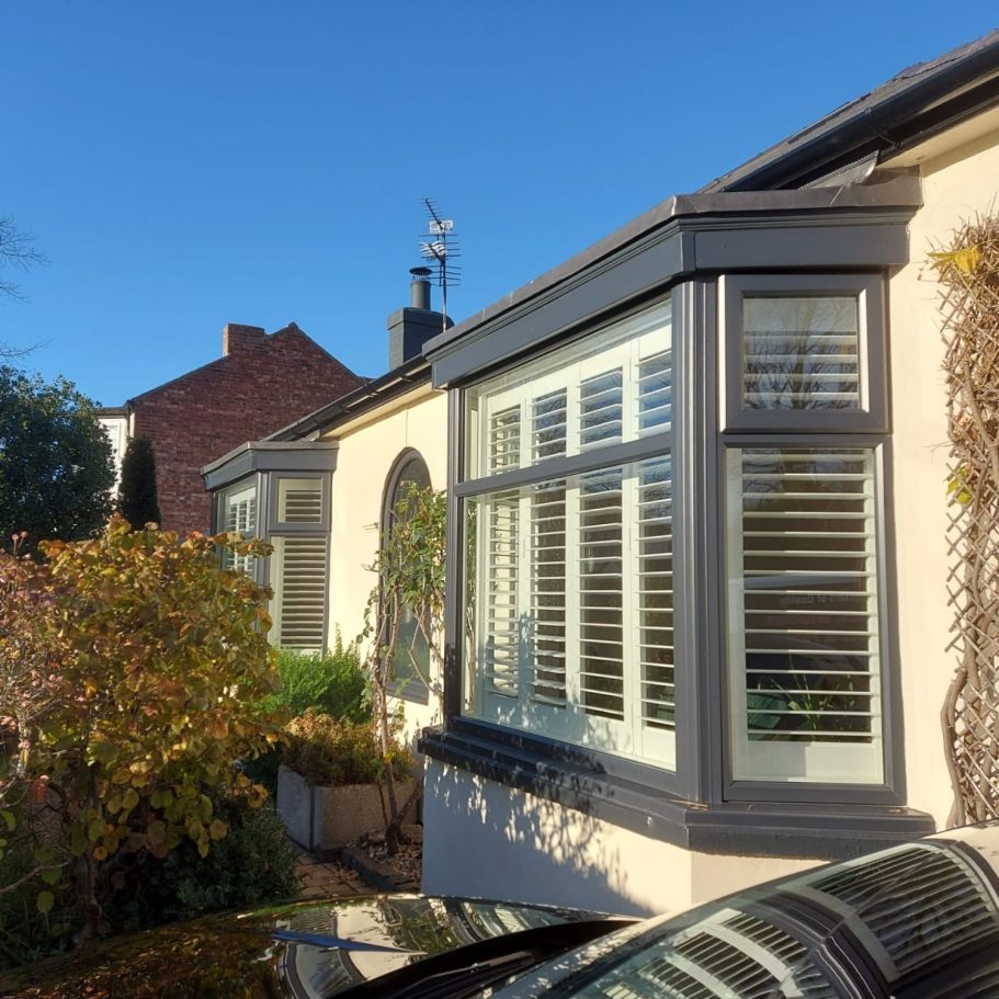 Close-up of a bungalow's bay window with shutters and surrounding foliage under a clear blue sky. near me