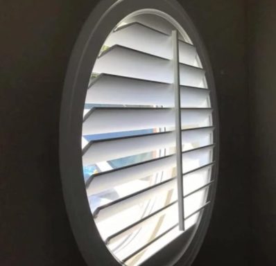 Oval window with wooden blinds partially open, revealing bright daylight outside.