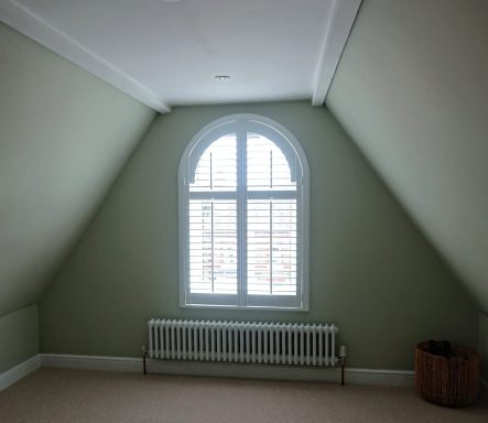 Empty attic room featuring a large arched window and a radiator against light green walls.
