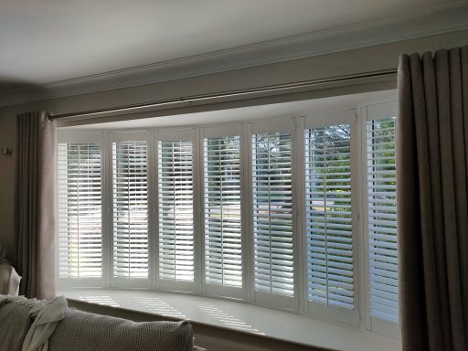 Bright interior with white plantation shutters and light curtains along a curved window.